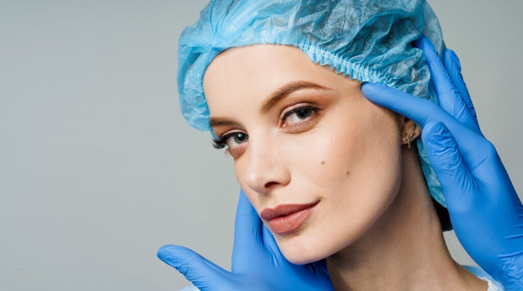 Mulher jovem com rosto maquiado, usando touca de cirurgia na cor azul. Há duas mãos, com luvas azuis, segurando seu rosto, possivelmente para mostrar quais procedimentos cirúrgicos podem ser realizado pelo consórcio de cirurgia plástica.